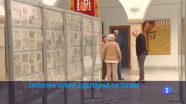 Carlos de las Heras, Remedios Tierno y otras personas conversando en la Asamblea de Extremadura. Se lee un rótulo que dice "Informe sobre apartheid en Israel".