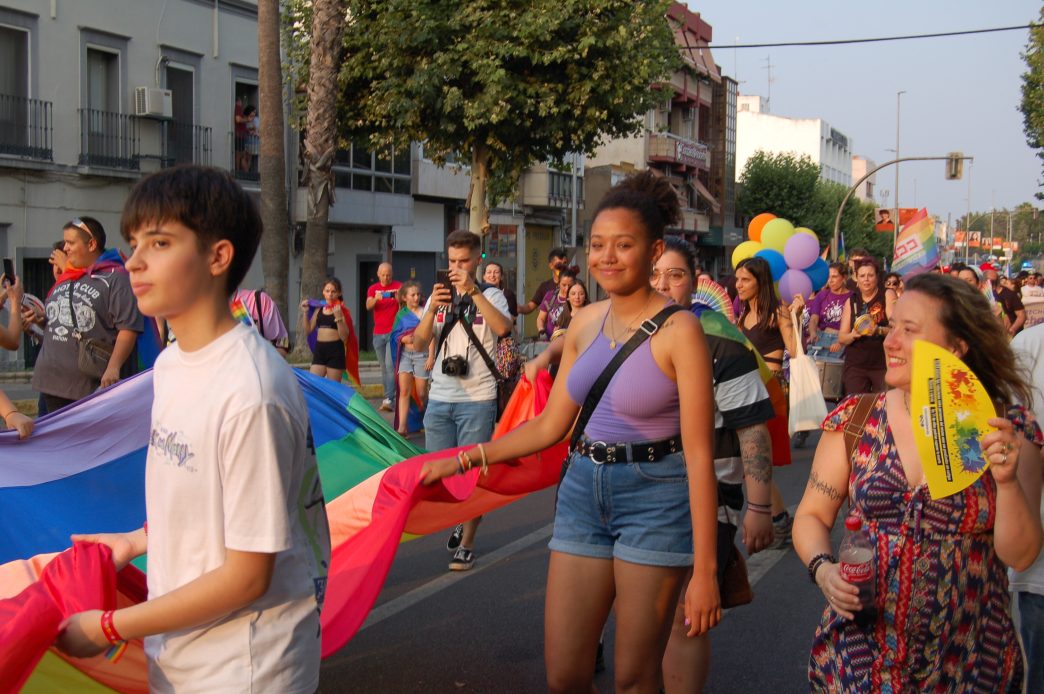Un grupo de personas portan una bandera arcoiris de varios metros de longitud. En el centro de la imagen una chica sonríe a la cámara