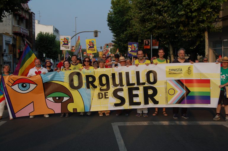 Activistas de Amnistía Internacional Extremadura tras una pancarta con el lema ORGULLO DE SER y la bandera inclusiva