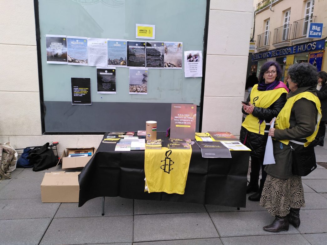 Dos activistas de Amnistía Internacional, con petos amarillos con el logo de la ONG, junto a una mesa con folletos y otros materiales. Al fondo, en los cristales, hay unos paneles en los que se intenta explicar el conflicto palestino-israelí.