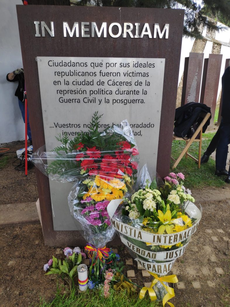Monumento con el rótulo IN MEMORIAN y una placa en la que puede leerse "Ciudadanos que por sus ideales republicanos fueron víctimas en la ciudad de Cáceres de la represión política durante la Guerra Civil y la posguerra." La parte inferior del texto no puede leerse por estar cubierta por dos ramos de flores, uno de los cuales tiene los colores de la bandera republicana, y el otro una cinta con el lema AMNISTÍA INTERNACIONAL VERDAD JUSTICIA REPARACIÓN.