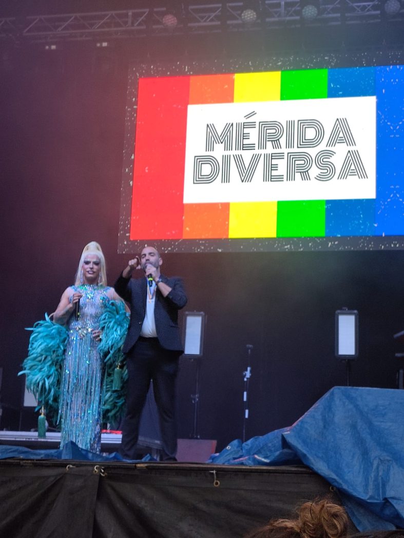 Una drag queen y su acompañante masculino presentando el evento desde el escenario. Al fondo, en la pantalla electrónica, puede leerse MÉRIDA DIVERSA sobre un fondo con los colores de la bandera arco iris.