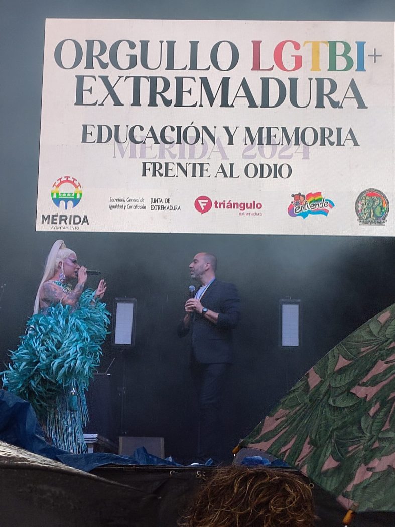 Otra instantánea de la pareja que presentaba el evento. En la pantalla electrónica puede leerse ORGULLO LGTBI+ EXTREMADURA, EDUCACIÓN Y MEMORIA FRENTE AL ODIO, y logos de el Ayuntamiento de Mérida, Junta de Extremaduira, Triángulo, Extremadura Entiende y Summa Diversitas