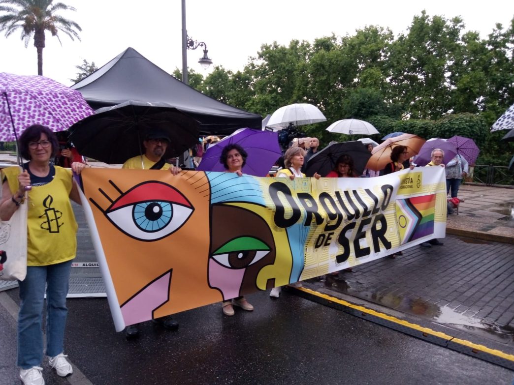 Activistas de AI portando una pancarta con el lema ORGULLO DE SER y la bandera LGTBIQ+ inclusiva.