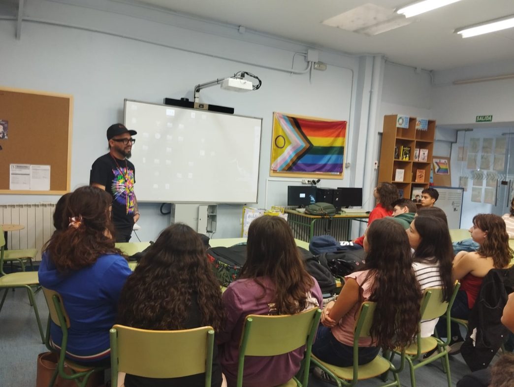 Yoelkis hablando al alumnado en un aula del IES Pérez Comendador. Tras él, junto a la pantalla, puede verse una bandera LGTBIQ+ inclusiva.