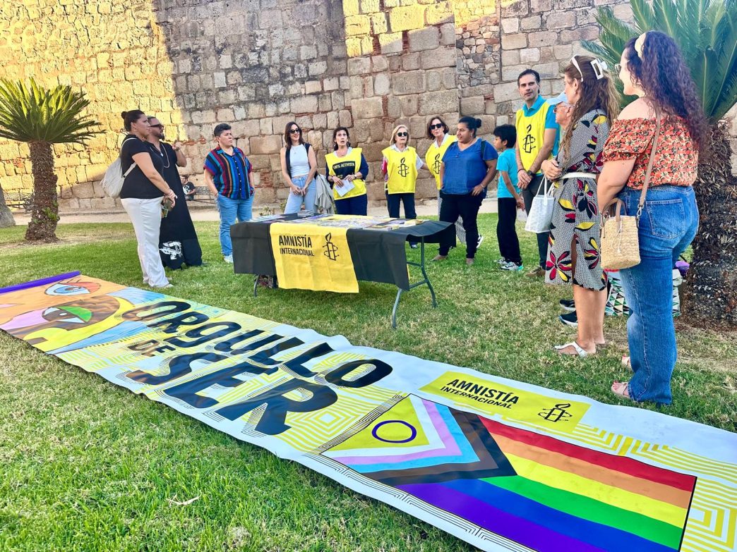 Yoelkis junto a varias personas de AI y otros colectivos en la Plaza de las Méridas del Mundo. En el suelo hay una pancarta de AI con el lema "Orgullo de ser". También hay una mesa con materiales de AI.