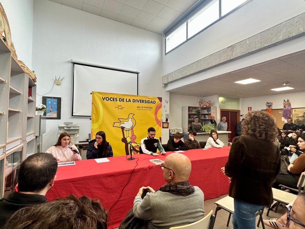 Portavoces del alumnado hablando en la bibilioteca al resto del centro. Están tras una mesa roja. Al fondo se ve un cartel del evento.