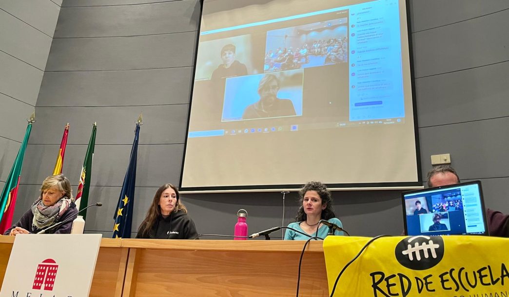 Varias personas sentadas tras una mesa. Sobre ellas, en una pantalla, se ven otras dos y el público asistente al acto.