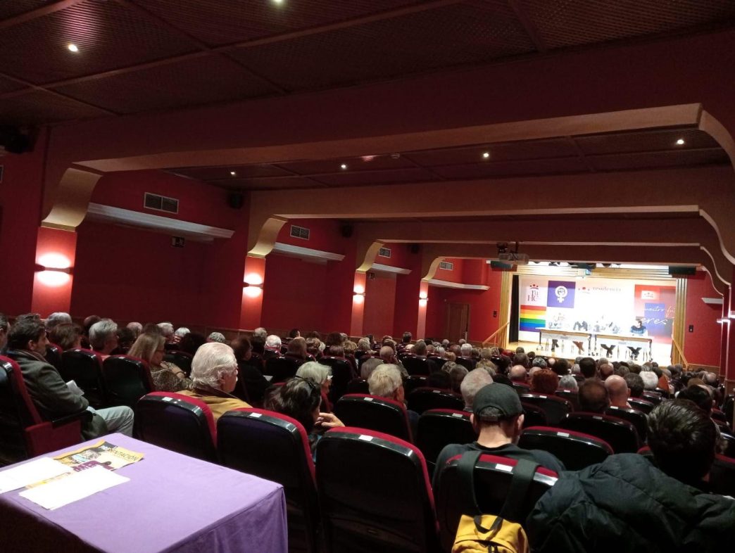 Salón de actos de la Residencia Universitaria Hernán Cortés de Badajoz. Puede verse abundante público y, en el escenario, una mesa con cuatro personas. Tras la mesa se ven una bandera arcoiris, un símbolo feminista, el logo de la residencia y otras imágenes.