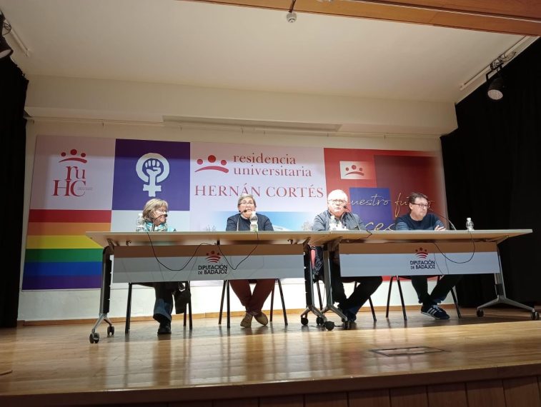 Escenario del Salón de Actos de la Residencia Hernán Cortés de Badajoz. Se ve una mesa con cuatro personas. Tras la mesa se ven una bandera arcoiris, un símbolo feminista y otras imágenes.