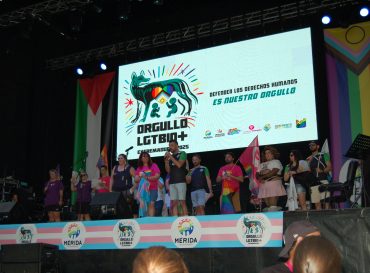 Representantes de las asociaciones organizadoras del Orgullo LGTBIQ+ Extremadura 2025 en un escenario, al final de la marcha. En la pantalla se lee el lema DEFENDER LOS DERECHOS HUMANOS ES NUESTRO ORGULLO.