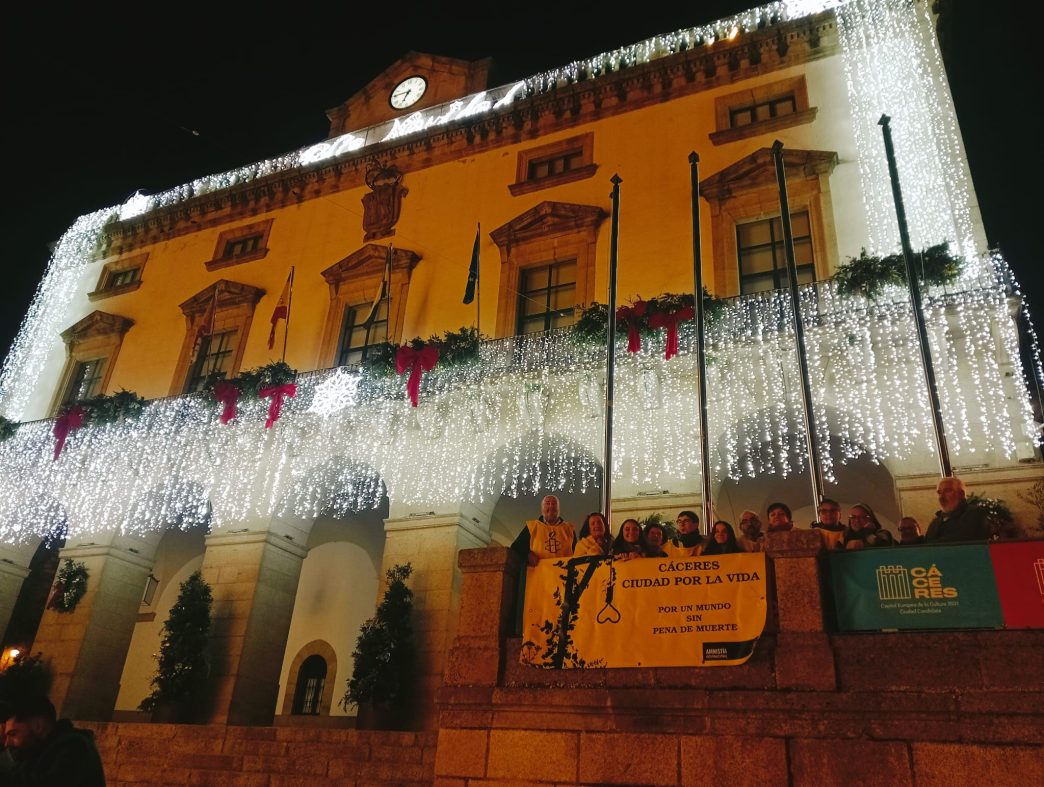 Un grupo de activistas de Amnistía Internacional con un cartel con el lema CÁCERES CIUDAD POR LA VIDA. De fondo se ve el Ayuntamiento de Cáceres, iluminado de amarillo y con decoración navideña.