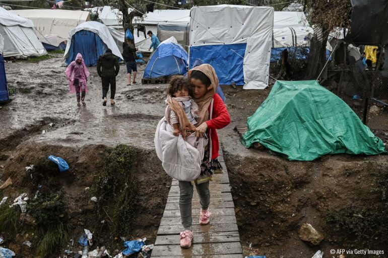 Niña con un bebé en brazos andando por un campo de refugiados en Grecia
