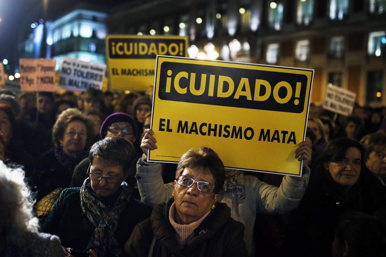 Una manifestación en el que se ve a una mujer mayor con cara triste y detrás una pancarta que reza "Cuidado, el machismo mata"
