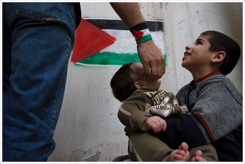 La mano de un hombre, con una bandera palestina en su muñeca, acaricia un bebé sujetado por un niño. En el fondo, a un metro, un pared con la bandera palestina pintada con spray.