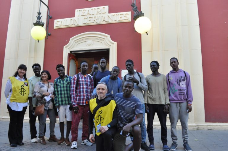 Fotografía de un grupo de jóvenes negros junto con dos miembros del equipo de AI Alcalá de Henares a la entrada del Teatro Salón Cervantes.